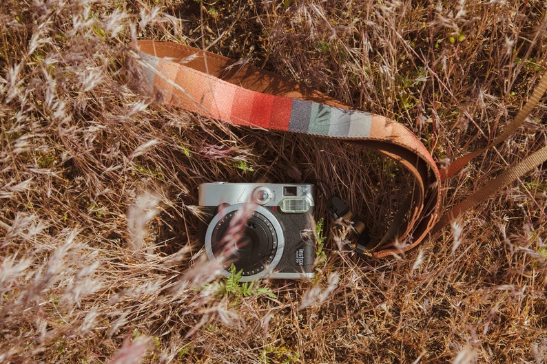 laying in the grass is a retro striped printed camera strap. Striped pattern of shades of red, orange, brown ,blue, and green. Camera connected to film camera.