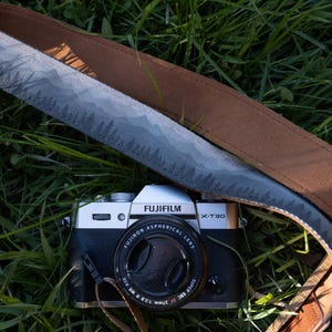 May include: A silver and black Fujifilm X-T30 camera with a Fujinon 27mm lens, featuring a camera strap with a grey mountain and forest landscape print on one side and a solid brown fabric backing, resting on a patch of green grass.
