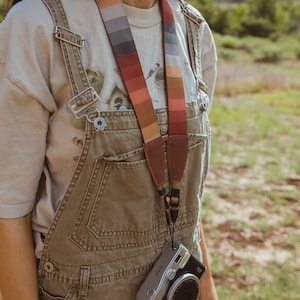sideview of women wearing around neck Retro striped printed camera strap. Striped pattern of shades of red, orange, brown ,blue, and green. Camera connected to film camera.