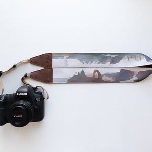 May include: A black Canon DSLR camera attached to a camera strap featuring a printed landscape design of a natural stone arch, evergreen trees, and mountains. The strap has brown leather end tabs and adjustable beige nylon webbing connectors.