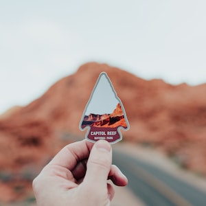 Op de afbeelding: Een sticker in de vorm van een pijlpunt met een afbeelding van een rode rotskloof en de tekst "Capitol Reef National Park".