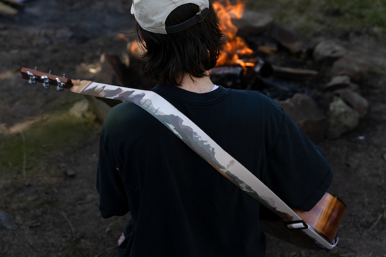 man sitting at campfire playing guitar wearing national park guitar strap in Color features 18 National Park illustrations printed along the length of the strap. Brown leather ends.