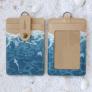 Op de afbeelding: Twee rechthoekige kaarthouders met een beige bovenzijde en een blauw gedeelte met oceaanthema, voorzien van silhouetten van zeeschildpadden, haaien en roggen. Eén houder heeft een bandje met een drukknoop en een icoon van een dennenboom.