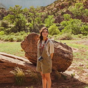women standing in valley in front of big rock wearing around neck Retro striped printed camera strap. Striped pattern of shades of red, orange, brown ,blue, and green. Camera connected to film camera