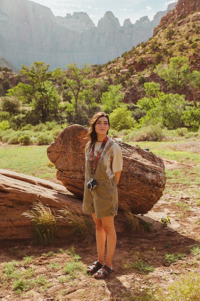 women standing in valley in front of big rock wearing around neck Retro striped printed camera strap. Striped pattern of shades of red, orange, brown ,blue, and green. Camera connected to film camera