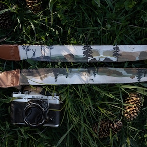 May include: A Fujifilm X-T30 camera with a black body and a lens cap attached to a long camera strap. The strap features a printed landscape design with silhouettes of pine trees, a deer, and mountains in shades of brown, green, and white.