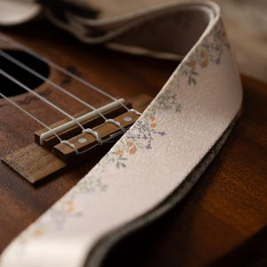 Cute tan floral strap printed with Little colored flowers across the length of the strap attached to Ukulele lying on wood floor