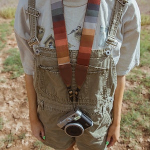 women wearing around neck Retro striped printed camera strap. Striped pattern of shades of red, orange, brown ,blue, and green. Camera connected to film camera.