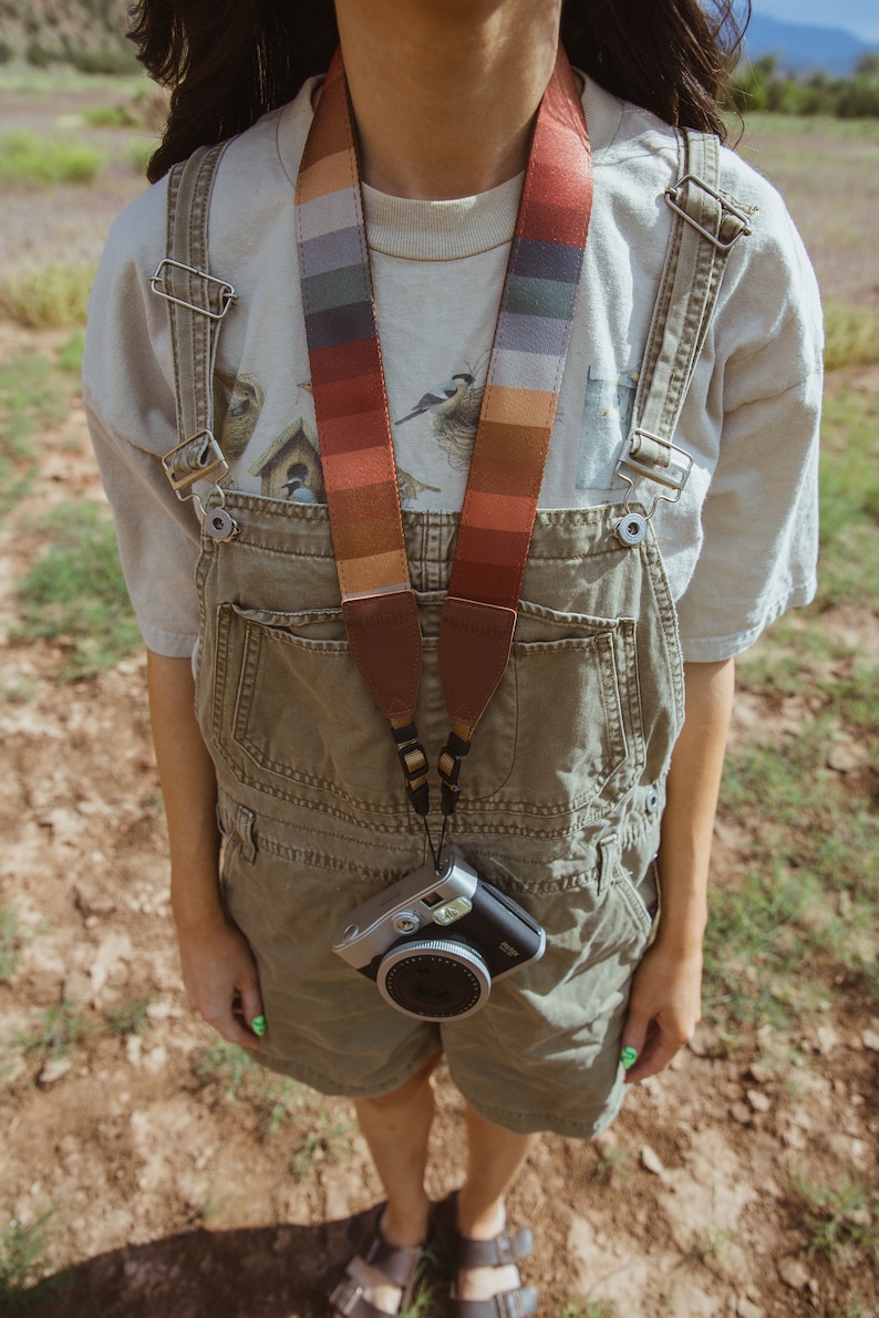 women wearing around neck Retro striped printed camera strap. Striped pattern of shades of red, orange, brown ,blue, and green. Camera connected to film camera.