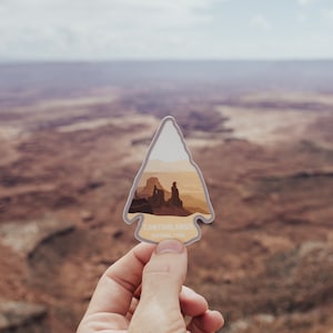 May include: A close-up of a Canyonlands National Park sticker in the shape of an arrowhead. The sticker is held in a hand against a backdrop of red rock canyons.