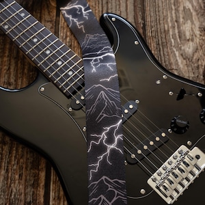 Könnte beinhalten: Eine schwarze E-Gitarre mit einer silbernen Brücke und einem schwarz-weißen Gitarrengurt mit einem Blitz- und Bergdesign.