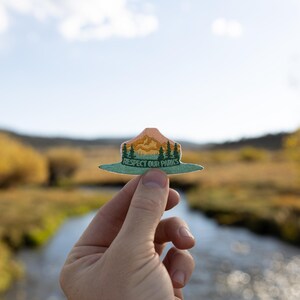 Hand holding out respect out parks park ranger hat patch in front of river landscape.