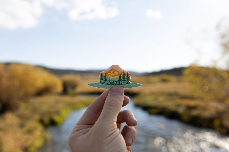 Hand holding out respect out parks park ranger hat patch in front of river landscape.