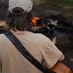 Man sitting in front of campfire playing acoustic guitar wearing a black guitar strap printed in black white white stars and dark gray mountains printed across the length of the strap
