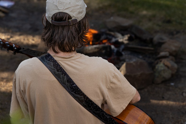 Man sitting in front of campfire playing acoustic guitar wearing a black guitar strap printed in black white white stars and dark gray mountains printed across the length of the strap