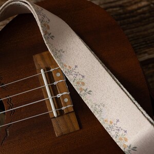 Cute tan floral strap printed with Little colored flowers across the length of the strap attached to Ukulele lying on wood floor