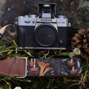 Pode incluir: Uma câmera Fujifilm X-T30 prateada com uma lente preta e uma alça de câmera de couro marrom com um padrão de cogumelos. A câmera está sentada em um leito de grama verde com pinhas.