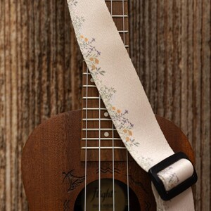 Cute tan floral strap printed with Little colored flowers across the length of the strap attached to Ukulele lying on wood floor