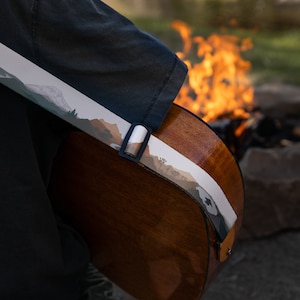 closeup of man sitting at campfire playing guitar wearing national park guitar strap in Color features 18 National Park illustrations printed along the length of the strap. Brown leather ends.