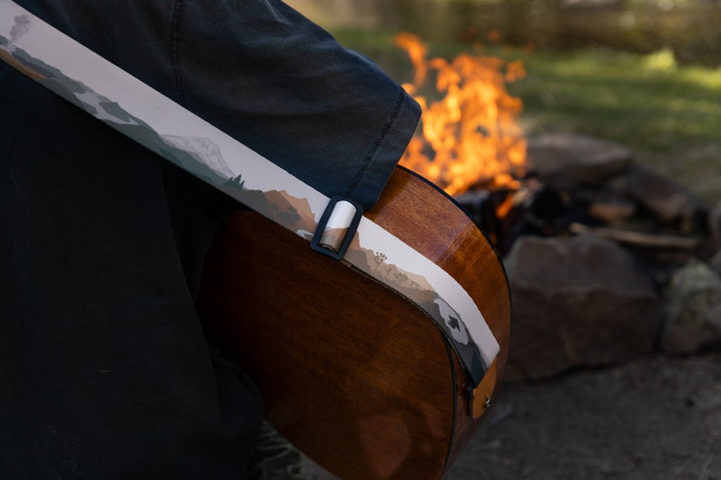 closeup of man sitting at campfire playing guitar wearing national park guitar strap in Color features 18 National Park illustrations printed along the length of the strap. Brown leather ends.