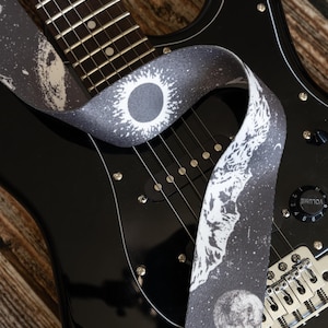 Puede incluir: Una guitarra eléctrica negra con una correa gris y blanca con un diseño celestial. La correa muestra imágenes del sol, la luna y las estrellas. La guitarra tiene un mando de volumen y herrajes plateados.