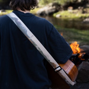 man sitting at campfire playing guitar wearing national park guitar strap in Color features 18 National Park illustrations printed along the length of the strap. Brown leather ends.