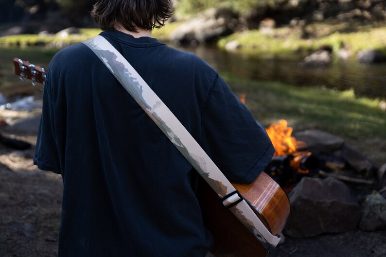 man sitting at campfire playing guitar wearing national park guitar strap in Color features 18 National Park illustrations printed along the length of the strap. Brown leather ends.