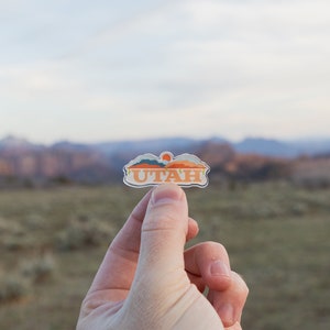May include: A clear sticker with a sunset scene and the word "UTAH" in orange letters. The sticker is being held in a hand against a blurred background of mountains and a cloudy sky.