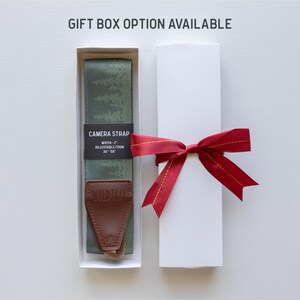 May include: A green and white camera strap with a brown leather adjustable strap and the text "CAMERA STRAP" and "WIDTH - 1" printed on a black label. The strap is in a white gift box with a red ribbon.