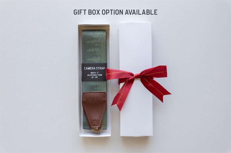 May include: A green and white camera strap with a brown leather adjustable strap and the text "CAMERA STRAP" and "WIDTH - 1" printed on a black label. The strap is in a white gift box with a red ribbon.