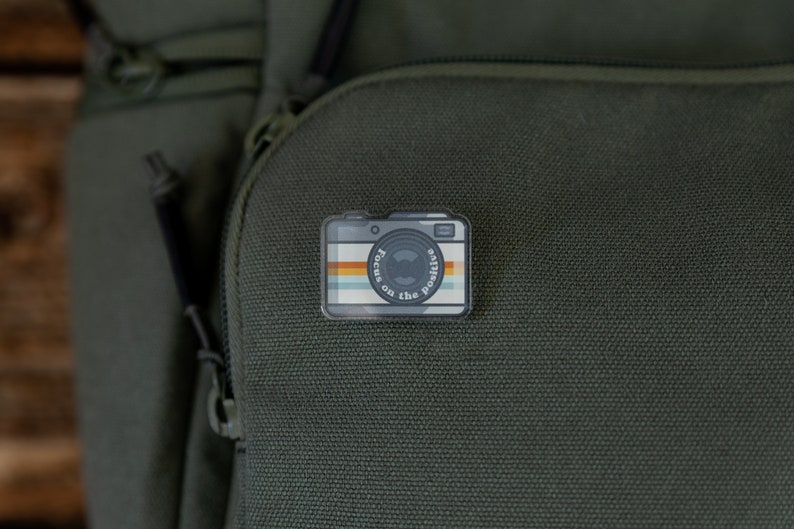 acrylic pin in the shape of a vintage camera with the words around the lens "Focus on the positive"