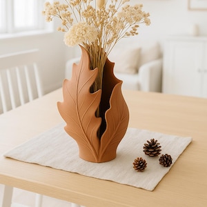 Könnte beinhalten: Eine terrakottafarbene Vase in Blattform, die getrocknete Blumen und Gräser enthält. Die Vase steht auf einem Holztisch mit einem neutralfarbenen Leinenläufer. Drei kleine Tannenzapfen sind in der Nähe der Vase angeordnet.