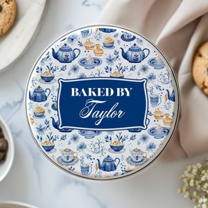 May include: A white round tin cookie container with a blue and gold floral pattern. The lid features a blue banner with the text "BAKED BY Taylor".