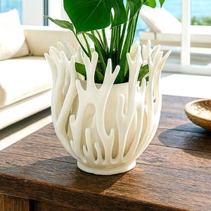 Peut inclure: Une plante Monstera verte luxuriante dans un pot en céramique blanc en forme de corail. La plante est posée sur une table en bois sombre, avec un canapé aux tons neutres et une vue sur la mer en arrière-plan. Un bol décoratif est également sur la table.