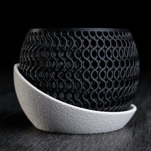 May include: A black, patterned planter pot with a matching white saucer. The pot has a repeating, wave-like design. The saucer is a simple, curved shape. The pot and saucer are sitting on a dark wooden surface.