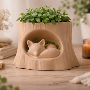 May include: A beige ceramic planter shaped like a tree stump, featuring a sleeping fox sculpture inside a hollowed-out section. The top of the planter is filled with green plants. A candle and other decorative items are in the background.