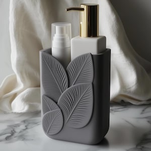Può includere: Un dispenser di sapone grigio con una pompa dorata e una bottiglia bianca, inserito in un supporto grigio con dettagli a foglie. Una bottiglia bianca con una pompa è anche nel supporto. Lo sfondo è un tessuto bianco.
