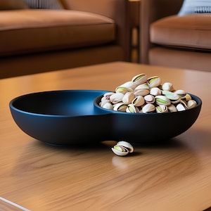 May include: A black, two-section serving bowl filled with pistachios. The bowl is on a wooden surface. The pistachios are open, showing the green nut inside. One pistachio is separate from the bowl.