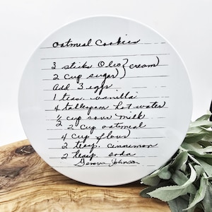 Może przedstawiać: Biały ceramiczny talerz z ręcznie napisanym przepisem na ciasteczka owsiane czarnym tuszem. Przepis zawiera składniki takie jak cukier, jajka i płatki owsiane. Talerz jest okrągły i leży na drewnianej powierzchni.