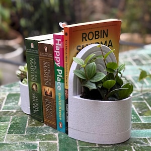 May include: A white bookend with a plant in it, holding up three books. The books are "The Monk Who Sold His Ferrari" by Robin Sharma, "Happy Place" by Emily Henry, and "A Clash of Kings" and "A Storm of Swords" by George R.R. Martin. The bookend is on a green tiled surface.