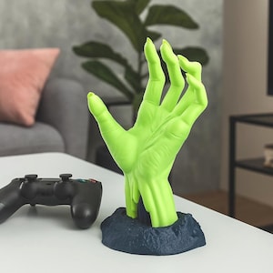Peut inclure: Une sculpture de main verte stylisée avec de longs ongles, émergeant d'une base bleu foncé. Une manette de jeu vidéo noire est au premier plan, suggérant un thème de jeu. La main est d'un vert vif.
