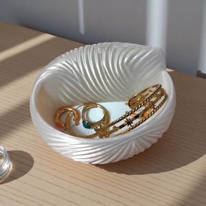 Könnte beinhalten: Eine weiße, muschelförmige Schmuckschale mit goldenen Ringen und Armbändern. Die Schale hat eine strukturierte, geriffelte Oberfläche. Der Schmuck umfasst einen Ring mit orangefarbenem Emaille-Design und einen Ring mit einem grünen Stein.