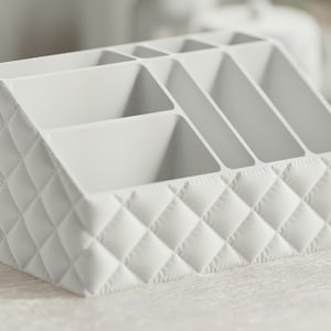 May include: A white, quilted organiser with multiple compartments. The organiser has a sloped design and is divided into several sections, ideal for storing makeup, office supplies, or other small items. The quilted pattern adds a touch of elegance.