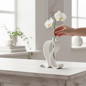 May include: A white, sculptural vase with a unique, flowing design. The vase holds a single stem of white orchids. The vase is on a white wooden table in a bright, airy room. The design is modern and minimalist.