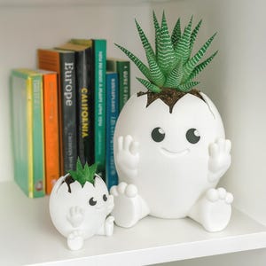 以下が含まれることがあります： 笑顔、腕、足が付いた、卵の殻の形をした2つの白いセラミックプランター。大きなプランターには緑色の多肉植物が入っています。同様のデザインの小さなプランターが隣にあります。背景には本があります。