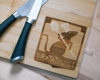 Puede incluir: Una tabla de cortar de madera con un diseño de rata chef y el texto "La Ratatouille". Dos cuchillos con mangos negros están sobre la tabla.