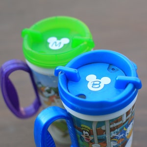 May include: Two colorful Mickey Mouse themed mugs with lids. The mug on the left has a green lid with a white Mickey Mouse head with the letter "M" inside. The mug on the right has a blue lid with a white Mickey Mouse head with the letter "B" inside.