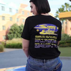 May include: A black t-shirt with a purple and yellow graphic design that says "Sammy Eclipse". The graphic design features a retro-style spaceship with stars. The text on the shirt reads "Bringing you the sunny side of entertainment from Planet Zor. Playing all your favorite hits. Gravity | Planetart | Boogie Space Angels | Down to Earth. Coming to a planet near you. The Starlight Stadium. Intergalactic Stadium. NASA's Colosseum. On tour."