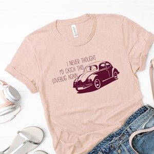 Pode incluir: Uma camiseta rosa claro com um gráfico vinho de um Volkswagen Beetle vintage e o texto "Nunca pensei que pegaria este fusca de novo."