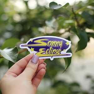 May include: A white sticker with a purple oval shape and the text "Sonny Eclipse" in yellow lettering. The oval is surrounded by a white outline and three yellow stars.